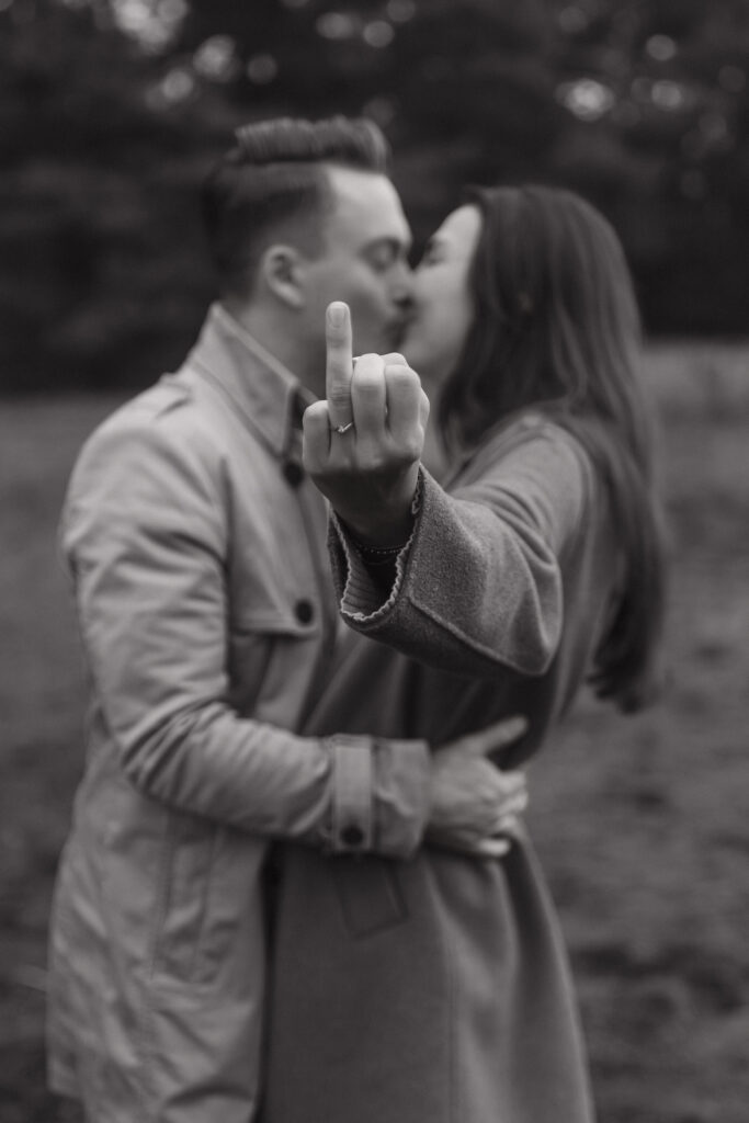 Verlobungsfotoshooting Frankfurt im Herbst. Es wird der Ringfinger mit Ring in die Kamera gehalten und im Hintergrund küsst sich das Paar.